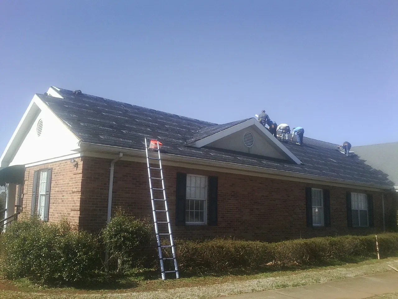 Crew tearing shingles off a brick ranch roof with a ladder set — Emergency Roof Repair work by our crew in Pavo