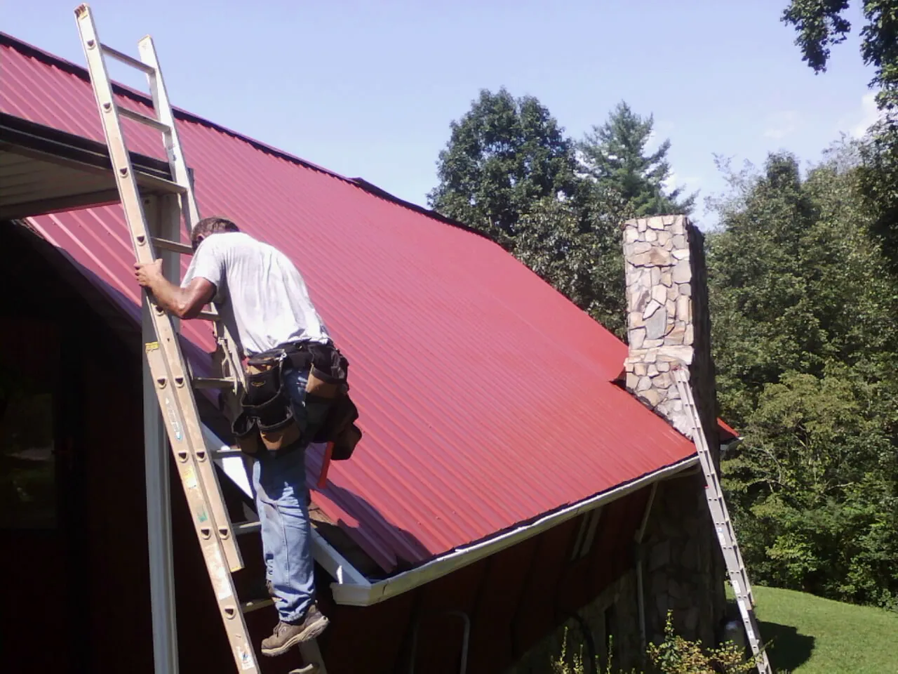 Roofer on a ladder beside a red metal roof and stone chimney — Soffit & Fascia Repair by Roofing Brothers in Pavo